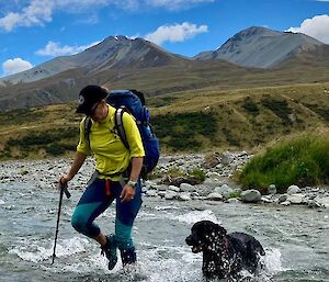 Billy travels over a shallow water creek with her dog Tui by her side. Large green mountainous landscape of New Zealand in the background