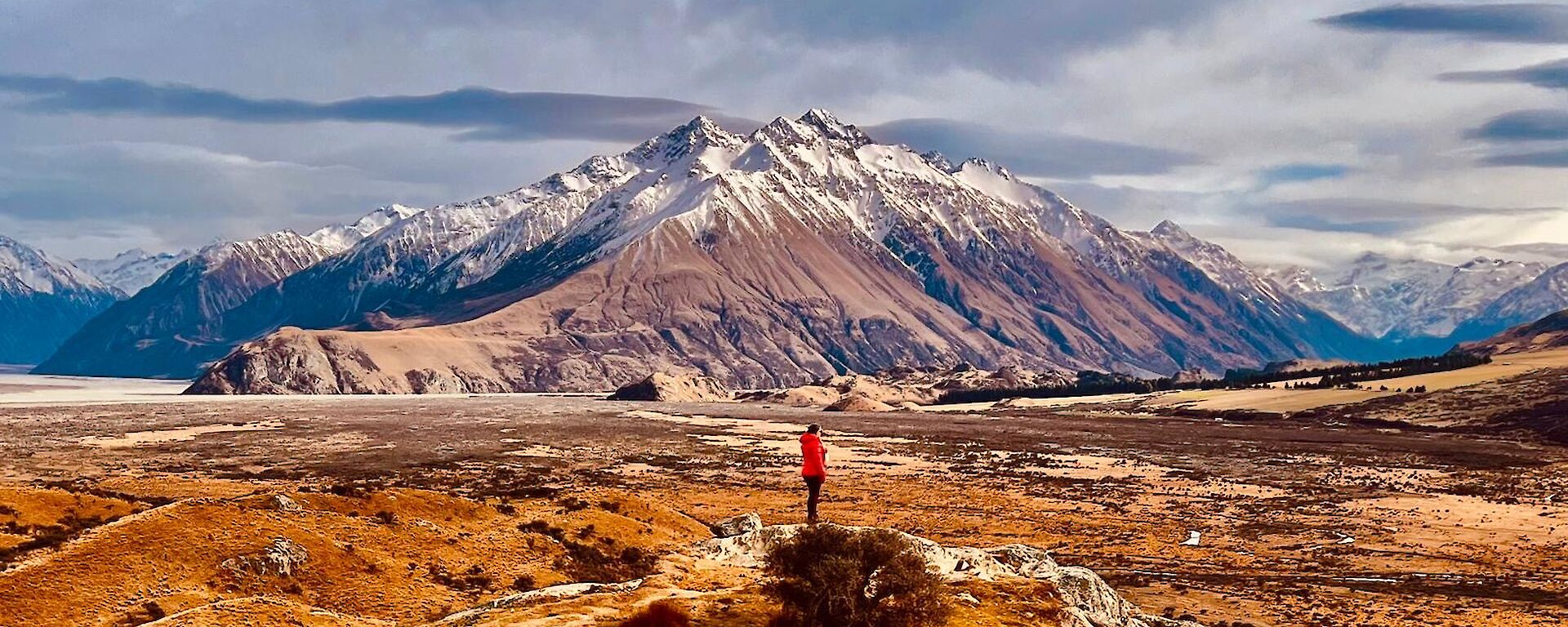 a very wide landscape in which Billy stands in an open plain leading into very large snow dusted mountains