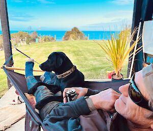 A photo of rob, Billy's' husband, and dog Tui relaxing on a hammock with ocean views in the distance