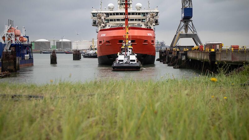 Looking across a grassy verge towards a large red ship being tugged out of dock