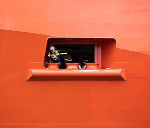 A close up of an open portal in the side of a red ship with a lady in hi-vis popping her head out