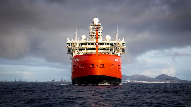 A large ship in the foreground with the hills, buildings and cranes of a small port on the horizon