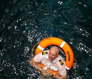 A man in a captains uniform and a life buoy over his head floating in some water