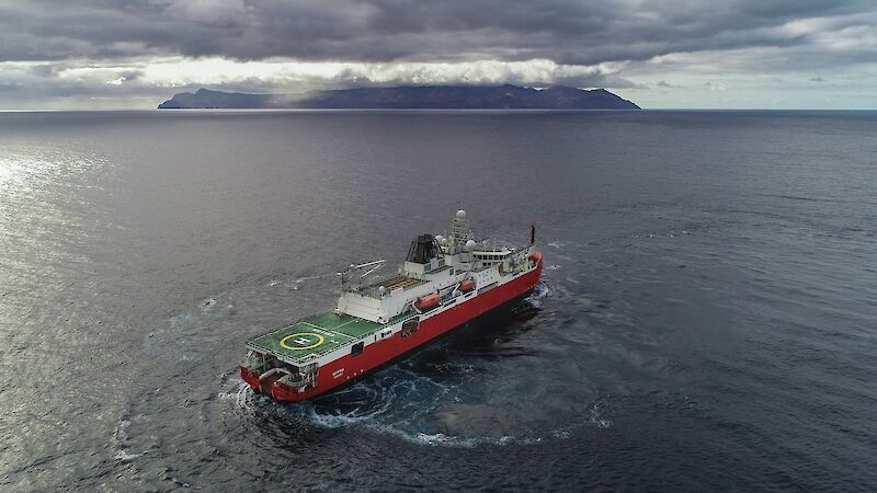 A ship in the ocean with an island visible in the distance. Circular trails can be seen in the water where the ship is turning