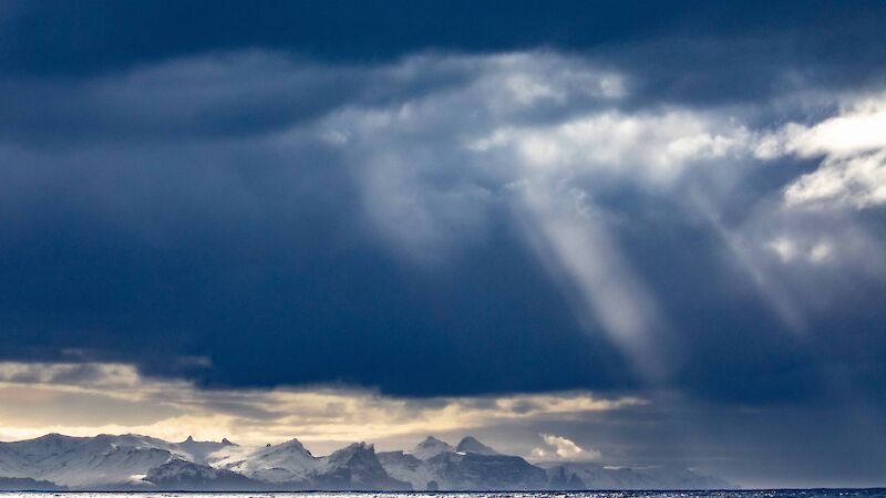 Sunlight streams through gathered dark clouds in the sky above a snow capped mountainous island.