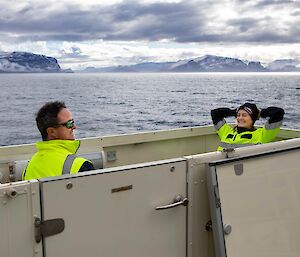 Two people in hi-vis sitting in a open top box at the back of a boat.  One person has their hands behind their head, leaning back and smiling.  Snowy mountains can be seen in the background.