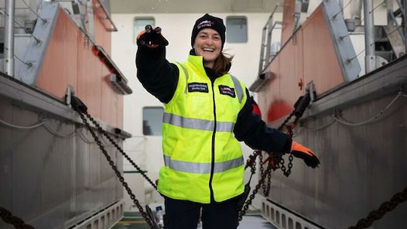 A woman in hi vis stands on the snowy dusted deck of a ship and throws a snowball at the camera