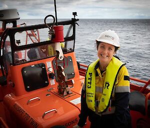 A woman in a hard hat and life jack sits on a barge at the side of a ship smiling to camera