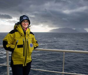 a woman stands at the rail of a ship in wet weather gear, smiling to camera, with an island visible on the horizon