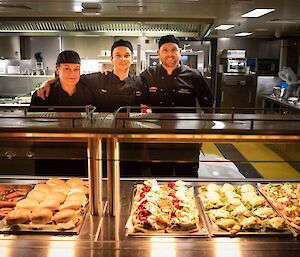 Three chefs stand arm in arm behind a bain marie of food, smiling to camera