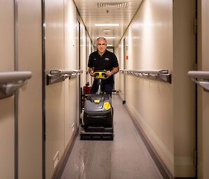 A man in a narrow ship corridor pushing a floor cleaning machine