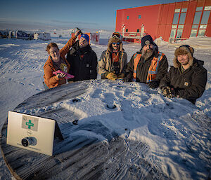 A woman holds a small ultrasound device to the head of a man, while three other men, all dressed in cold weather clothes look on. The team are standing around a wagon wheel table looking at a laptop that shows the ultrasound imagery.