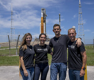Two men and two women in black t-shirts and jeans standing in front of a distant space rocket.