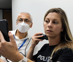 A woman using a small, hand-held ultrasound and mobile phone to look at her jugular vein, while a doctor in a mask looks on.