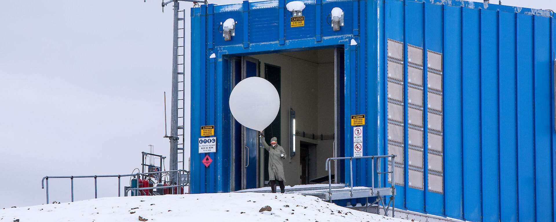 A man holds a white, hydrogen-filled balloon above his head, standing on snow in front of a blue building.