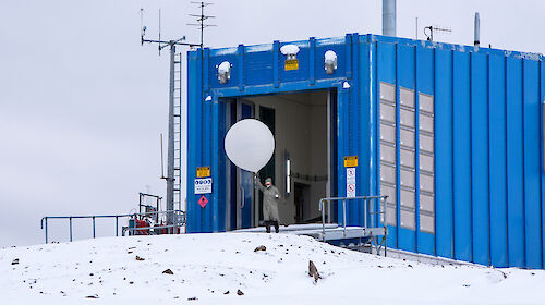 A man holds a white, hydrogen-filled balloon above his head, standing on snow in front of a blue building.
