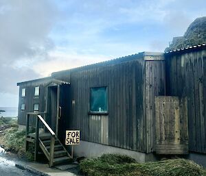 A grey weatherboard building with a 'for sale' sign out the front