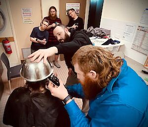 an expeditoner hair being cut, one using the clippers, one holding a bowl atop the head and three engrossed on-lookers