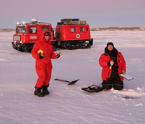 Two people in red outdoor gear standing on the ice in front of a hole, with a red Hägglunds behind them