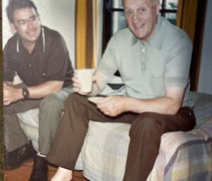Two men sit together on single bed, one holding a mug, smile to camera