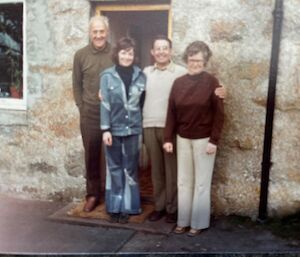 Group of four people stand on stoop of front door in front of stone house