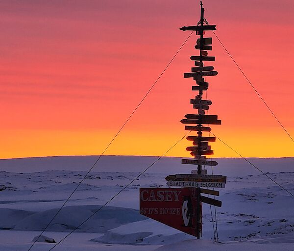 A portrait image depicting the Casey sign low lit in the foreground of backdropped background of vibrant sun arrays contrasted against a snow filled rocky outcrop