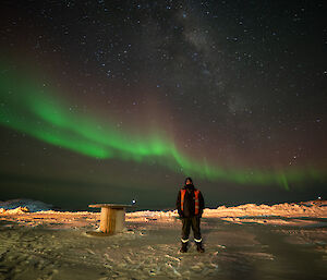 An expeditioner stands in front of a green and pink aurora in the night sky