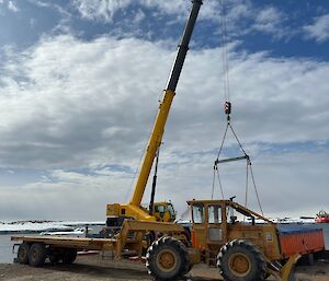 A large flat bedding truck awaits its next load as a crane swings container atop its bed, an image down by the wharf where supplies come off the ship