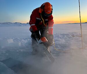 an image of James using a chain saw submerged into the water as he cuts away a portion of sea ice. a spectacular sunrise is dawning