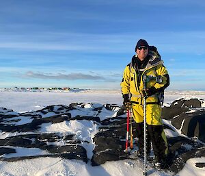 A man in yellow outdoor gear standing on a rocky and snowy hill with blue sky and snow in the background