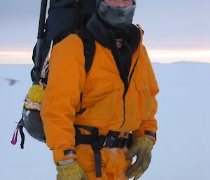 A man in orange outdoor gear and face muff stands on the snow