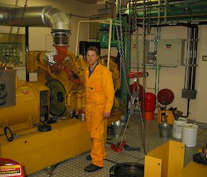 A young man stands in a mechanical workshop dressed in an orange worksuit