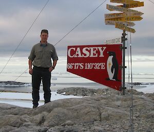 A man stands on a rocky slope with snow in the background, next to a sign that says Casey
