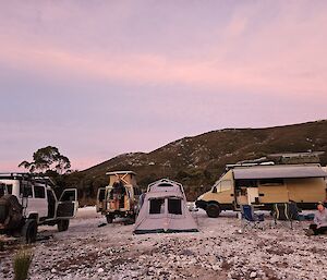 Three camper vehicles and a tent set up on white quartzite rock-covered ground with a mountain in the distance