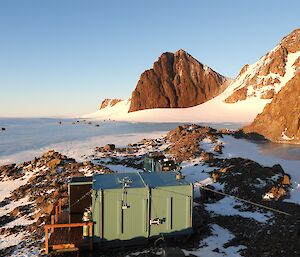 A green shipping-container hut in a picturesque snow-covered location surrounded by rocky mountain peaks