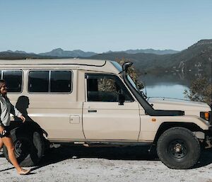A woman walks beside a Toyota troop carrier with a lake and mountains behind it.