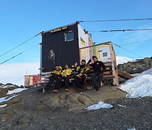 A group of people sit in front of a hut with a shed behind them and rock and snoe