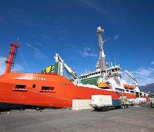 A large red ship tied up beside a wharf with shipping containers being loaded in to its cargo deck by a crane