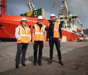 Three people in orange high-vis vests stand beside a red ship