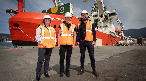Three people in orange high-vis vests stand beside a red ship