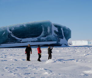 Three expeditioners on the sea ice taking photos of a magnificent blue/green coloured iceberg or jade berg
