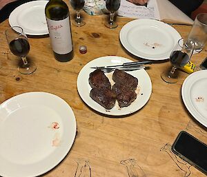 Round wooden table with table settings, serving plate of steak in the middle, bottle of wine with glasses