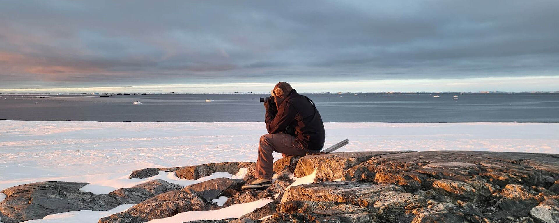 an expeditioner sits atop a rocky outcrop capturing the sunset through his camera