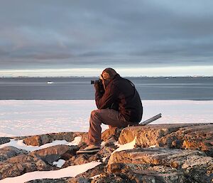 an expeditioner sits atop a rocky outcrop capturing the sunset through his camera