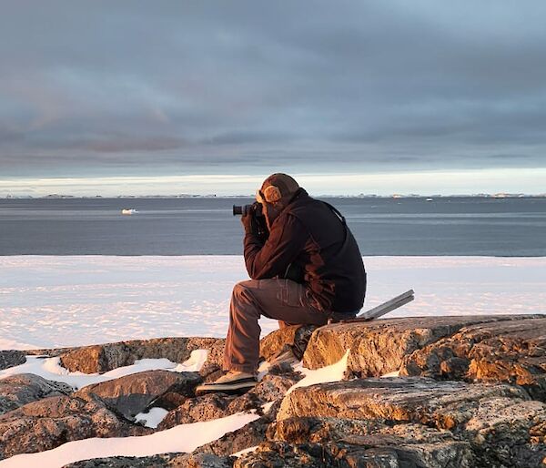 an expeditioner sits atop a rocky outcrop capturing the sunset through his camera