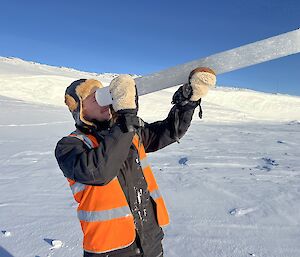 A man in hi vis stands on a snowy slope, holding an ice core to his face like a telescope
