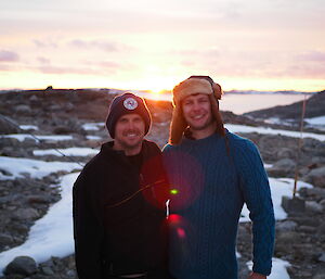 two expeditioners stand in the foreground of a view of  a vibrant sunset, with facial expressions of joy