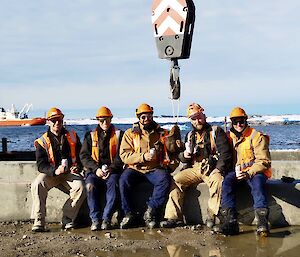 5 expeditioners proudly sit at the wharf where a crane hook plunging into the foreground of the image, the Resupply vessel postures in the bay in the backround