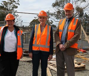 Two women and one man in orange high vis, stand near a metal fence on a building site