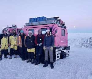 A group of people standing on a snowy slope with a pink snow tractor